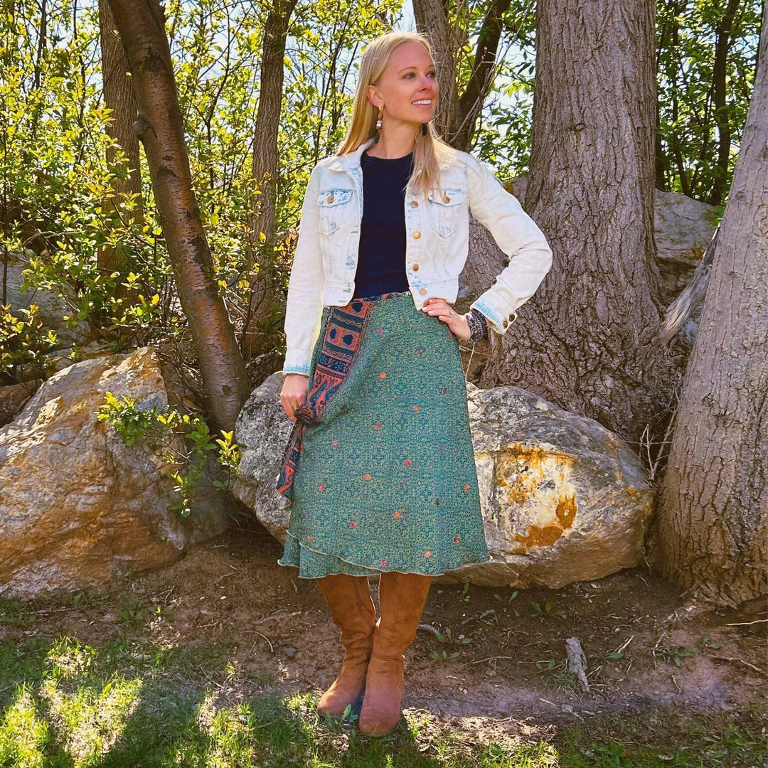 Woman outdoors in denim jacket, navy top, green skirt, and brown boots, with long blonde hair.