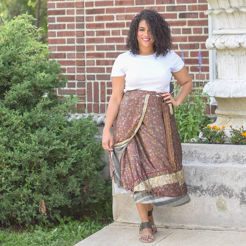 A tanned woman with curly hair is standing outside a brick and concrete building. She's wearing a white tee-shirt, sandals, and a floral brown sari wrap skirt.