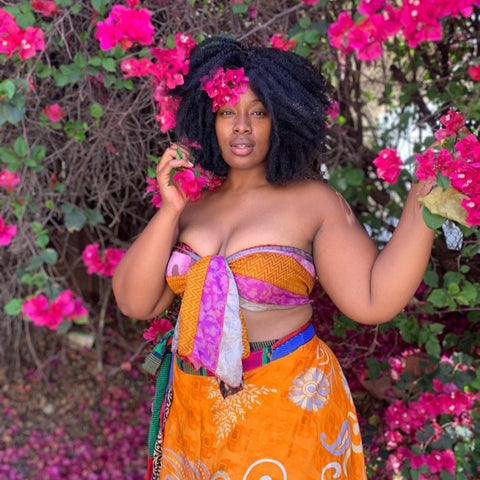 A gorgeous woman with a large afro, a pink and orange twist top, and an orange sari wrap skirt, is posing underneath a flowering bush.