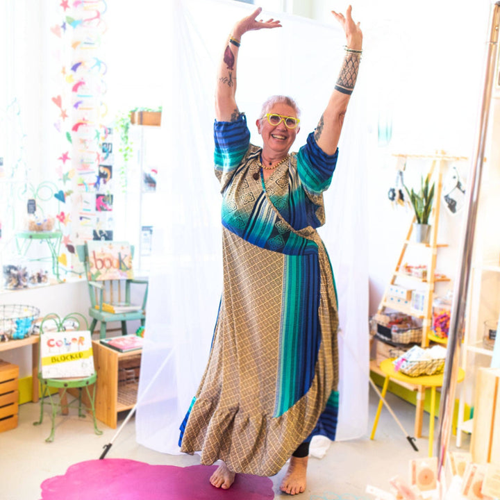 Woman in a colorful dress and yellow glasses posing joyfully in a bright, eclectic room.